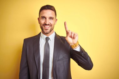 Sarı izole arka plan üzerinde genç yakışıklı iş adamı gösteren ve kendinden emin ve mutlu gülümseyerek parmak bir numara ile işaret.