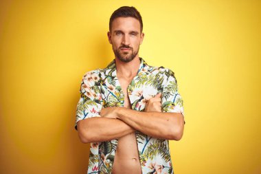 Yakışıklı adam sarı izole arka plan şüpheci ve sinir üzerinde yaz hawaii çiçek gömlek giyen, çapraz kolları ile yüzünde onaylamayan ifade. Negatif kişi.