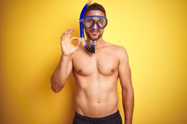 Genç yakışıklı gömleksiz adam izole sarı arka plan üzerinde dalış gözlüğü giyen parmakları ile ok işareti yapıyor, mükemmel sembolü