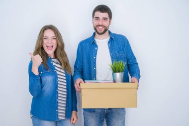 Genç çift tutarak karton kutu beyaz izole arka plan üzerinde yeni eve hareket eden büyük bir gülümseme ok işareti yapıyor mutlu, parmakları ile başparmak, mükemmel işareti