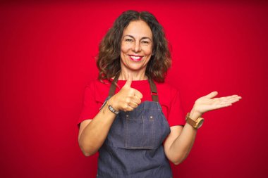 Orta yaş yaşlı kadın kırmızı izole arka plan üzerinde önlük üniforma giyen palmiye el gösteren ve başparmak ile ok jest yapıyor, mutlu ve neşeli gülümseyerek