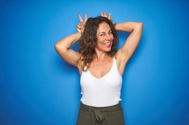 Kıvırcık saçlı orta yaş yaşlı kadın mavi izole arka plan üzerinde duran komik ve tavşan kulakları gibi baş parmakları ile deli poz, neşeli gülümseyen