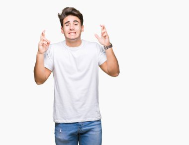 Umut ve gözleri kapalı geçiş parmaklarıyla gülümseyen izole arka plan üzerinde beyaz t-shirt giyen genç yakışıklı adam. Şans ve batıl kavramı.