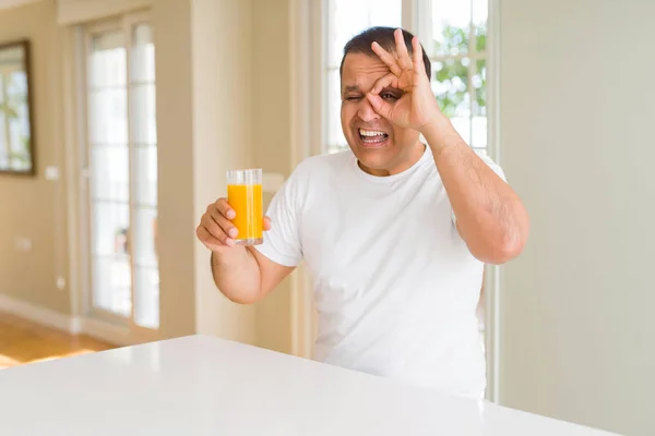 自宅でオレンジジュースのグラスを飲む中年の男は 指を通して見て目に手でOkサインをやって笑顔で幸せな顔をしています — ストック写真