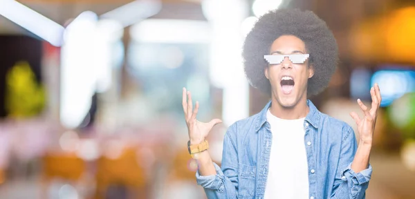 Young african american man with afro hair wearing thug life glasses crazy and mad shouting and yelling with aggressive expression and arms raised. Frustration concept.