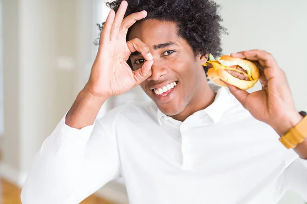 African American hungry man eating hamburger for lunch with happy face smiling doing ok sign with hand on eye looking through fingers