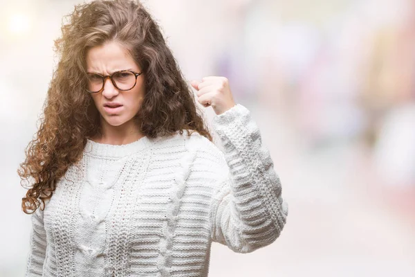 Beautiful brunette curly hair young girl wearing winter sweater over isolated background angry and mad raising fist frustrated and furious while shouting with anger. Rage and aggressive concept.