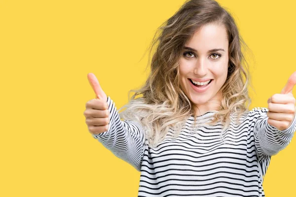Beautiful young blonde woman wearing stripes sweater over isolated background approving doing positive gesture with hand, thumbs up smiling and happy for success. Looking at the camera, winner gesture.