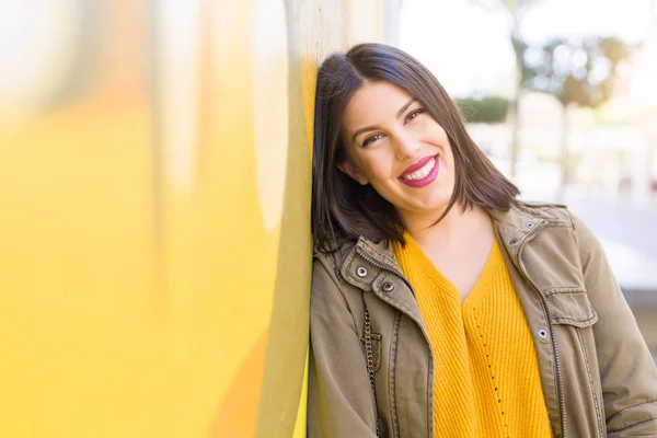 Beautiful young woman smiling confident and cheerful leaning on yellow wall, walking on the street of the city on a sunny day