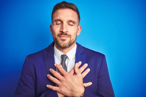 Close up picture of young handsome business man over blue isolated background smiling with hands on chest with closed eyes and grateful gesture on face. Health concept.