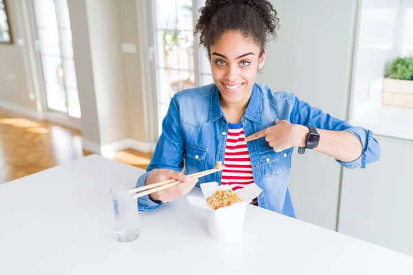Young african american woman eating asian noodles from delivery box with surprise face pointing finger to himself