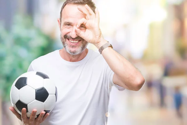 孤立した背景に指を通して見る目に手で サインをして笑っている幸せそうな顔でサッカー サッカー ボールを保持している中年白髪年配の男性 — ストック写真