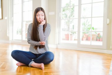 Güzel Asyalı kadın evde yerde yalınayak oturan stresli ve ağız tırnakısırma elleri ile sinirli görünümlü. Anksiyete sorunu.