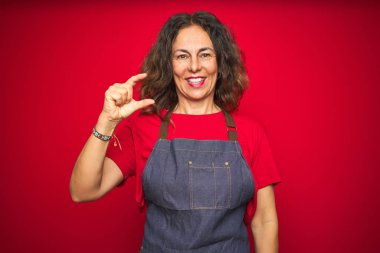 Orta yaş yaşlı kadın kırmızı izole arka plan üzerinde önlük üniforma giyen gülümseyen ve emin el parmakları görünümlü ve kamera ile küçük boyutlu işareti yapıyor gesturing. Ölçü kavramı.