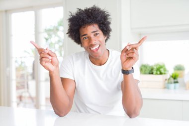 Genç afro-amerikan adam evde oturan rahat beyaz t-shirt giyen farklı yönlere parmakları ile işaret güvenerek gülümseyerek. Reklam için kopya alanı