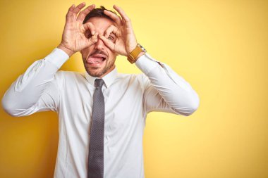 Genç yakışıklı iş adamı sarı izole arka plan üzerinde zarif beyaz gömlek giyen dürbün dil dışarı yapışmasını gibi ok jest yapıyor, gözleri parmaklardan bakarak. Çılgın ifade.
