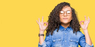 Gözlük sakin ol kıvırcık saçlı Genç güzel kadın ve gülümseyen gözlerle parmak ile meditasyon jest yapıyor kapalı. Yoga kavramı.