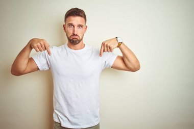 Genç yakışıklı adam izole arka plan üzerinde rahat beyaz t-shirt giyen üzgün ve üzgün görünümlü aşağı işaret, parmakları ile yönünü gösteren, mutsuz ve depresif.