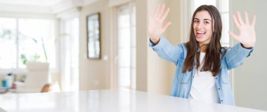 Evde beyaz masada oturan ve kendinden emin ve mutlu gülümserken parmaklarıyla on numara işaret eden güzel genç kadının geniş açılı resmi.