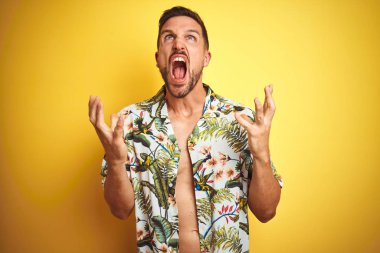 Yakışıklı adam sarı izole arka plan deli ve deli bağırarak ve agresif ifade ve kollar kaldırdı ile bağırıyor üzerinde yaz hawaii çiçek gömlek giyiyor. Hayal kırıklığı kavramı.