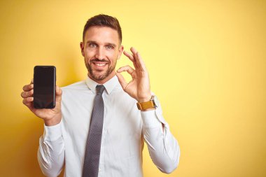 Genç yakışıklı iş adamı sarı izole arka plan üzerinde parmaklar, mükemmel sembolü ile ok işareti yapıyor akıllı telefon ekranı gösteren