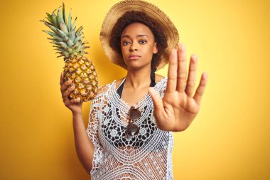 Afro saçlı genç afro-amerikan kadın ciddi ve kendinden emin ifade, savunma jesti ile durdurmak işareti yapıyor açık el ile sarı izole arka plan üzerinde pineaple tutan