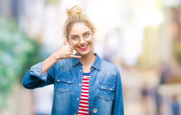 Young beautiful blonde woman wearing glasses over isolated background smiling doing phone gesture with hand and fingers like talking on the telephone. Communicating concepts.