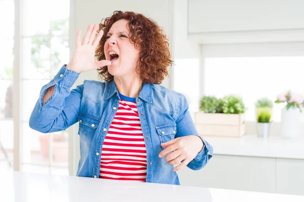 Evde kot ceket giyen kıvırcık saçlı orta yaş yaşlı kadın bağırarak ve ağızda el ile yan yüksek sesle bağırıyor. İletişim kavramı.