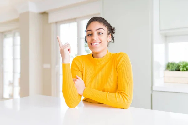 Beautiful african american woman with afro hair wearing a casual yellow sweater with a big smile on face, pointing with hand finger to the side looking at the camera.