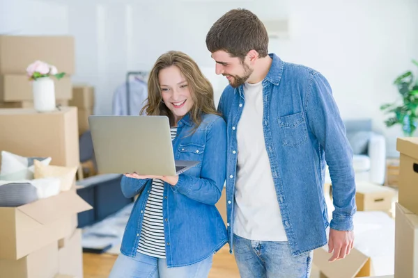 Young couple using computer laptop standing on a room around cardboard boxes, happy for moving to a new apartment