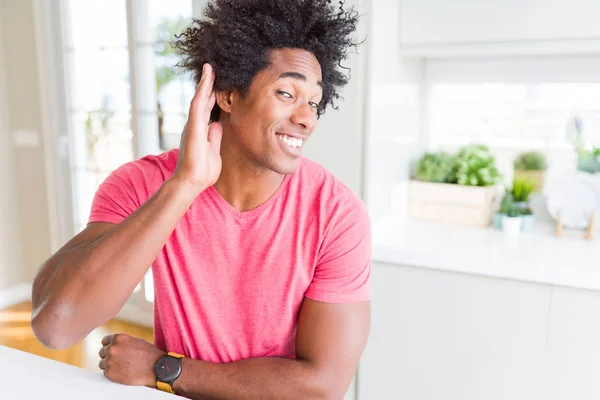 African American business man wearing pink casual t-shirt smiling with hand over ear listening an hearing to rumor or gossip. Deafness concept.