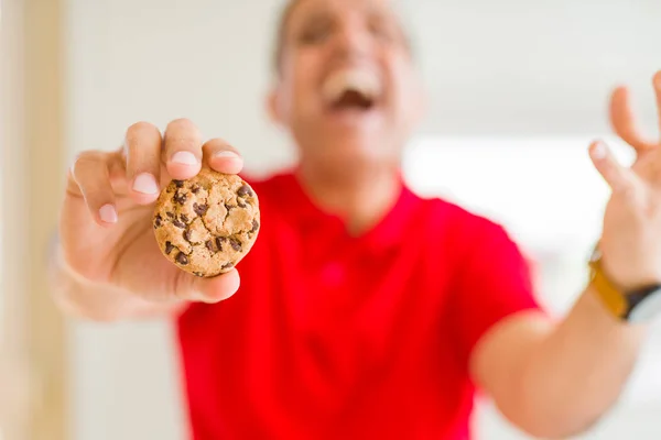 Middle age man eating chocolate chips cookies at home very happy and excited, winner expression celebrating victory screaming with big smile and raised hands