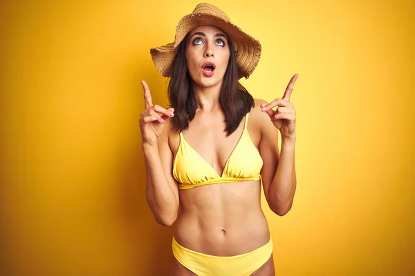穿着黄色比基尼和夏季帽子在孤立的黄色背景的美丽女人惊讶和惊讶抬头 用手指和抬起手臂 — 图库照片