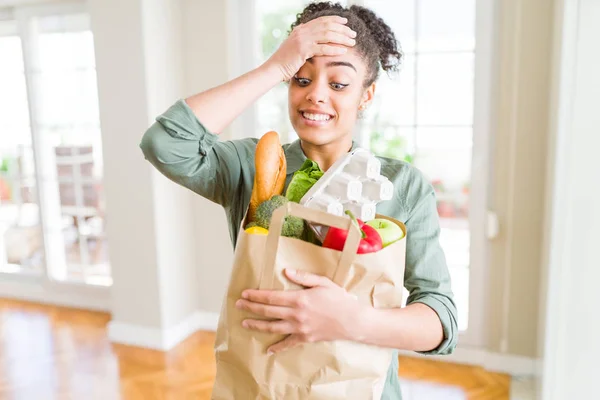 Young african american girl holding paper bag of groceries from supermarket stressed with hand on head, shocked with shame and surprise face, angry and frustrated. Fear and upset for mistake.