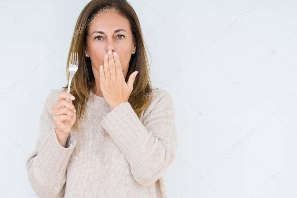 Middle age woman holding silver fork metal over isolated background cover mouth with hand shocked with shame for mistake, expression of fear, scared in silence, secret concept