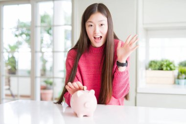 Güzel Asyalı kadın çok mutlu ve heyecanlı piggy çanta içinde bir sikke koyarak, kazanan ifade büyük bir gülümseme ile çığlık zafer kutluyor ve ellerini kaldırdı