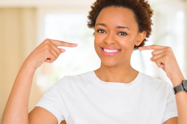 Genç güzel afro-amerikan kadın rahat beyaz t-shirt giyen yüzünde gülümseme ile emin görünümlü, parmakları gururlu ve mutlu kendini işaret.