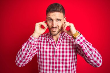 Yüksek sesle müzik sesi için rahatsız ifade ile parmakları ile kulakları kapsayan kırmızı izole arka plan üzerinde Genç yakışıklı adam. Sağır kavramı.