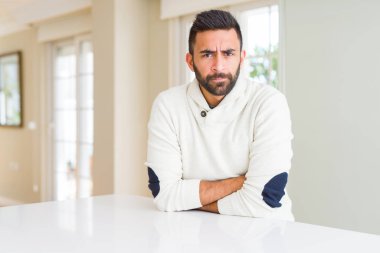 Yakışıklı İspanyol adam evde rahat beyaz kazak giyen şüpheci ve sinirli, çapraz kolları ile yüzünde onaylamayan ifade. Negatif kişi.
