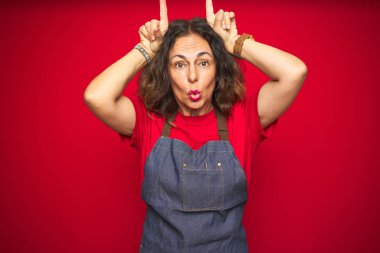 Orta yaş yaşlı kadın kırmızı izole arka plan üzerinde önlük üniforma giyen boğa boynuzları gibi baş üzerinde parmak ile komik jest yapıyor