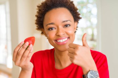 Genç afro-amerikan kadın kahvaltı için taze çilek yeme büyük bir gülümseme ok işareti yapıyor mutlu, parmakları ile başparmak, mükemmel işareti