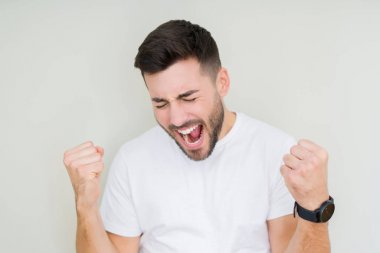 Gülümseyen ve başarı için çığlık izole arka plan çok mutlu ve heyecanlı kaldırdı, silah ile kazanan jest yapıyor üzerinde rahat beyaz t-shirt giyen genç yakışıklı adam. Kutlama kavramı.