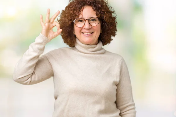 美しい中間エイガー タートルネックのセーターと手と指で サインをしている肯定的な孤立した背景を浮かべて上ガラスを身に着けている年配の女性 成功した式 — ストック写真