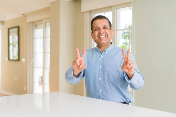Evde Oturan Orta Yaşlı Adam Gülümseyerek Kameraya Bakarak Parmaklarını Göstererek — Stok fotoğraf