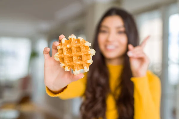 Young woman eating sweet waffle pastry surprised with an idea or question pointing finger with happy face, number one