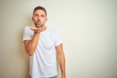 Bir öpücük el ile güzel ve seksi olmak hava üfleme kamera bakarak izole arka plan üzerinde rahat beyaz t-shirt giyen genç yakışıklı adam. Aşk ifade.