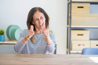 Orta yaş yaşlı kadın evde masada oturan mutlu ve komik yüzü ile kameraya parmaklarını işaret. İyi enerji ve titreşimler.