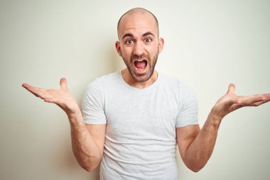 Sakallı genç kel adam izole arka plan üzerinde rahat beyaz t-shirt giyen deli kutluyor ve kollarını kaldırdı ve açık gözleri heyecan çığlık ile başarı için hayran. Kazanan kavramı