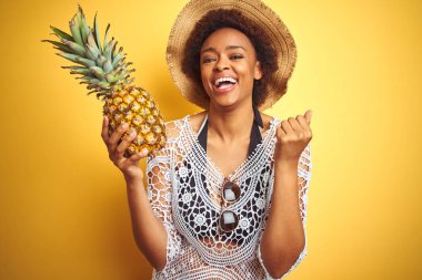 Afro saçlı genç afro-amerikan kadın sarı izole arka plan üzerinde pineaple tutan gurur çığlık ve zafer ve başarı çok heyecanlı kutluyor, duygu tezahürat