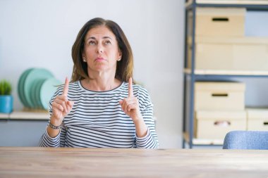 Orta yaş yaşlı kadın evde masada oturan üzgün ve üzgün görünümlü kadar işaret, parmakları ile yön gösteren, mutsuz ve depresif.